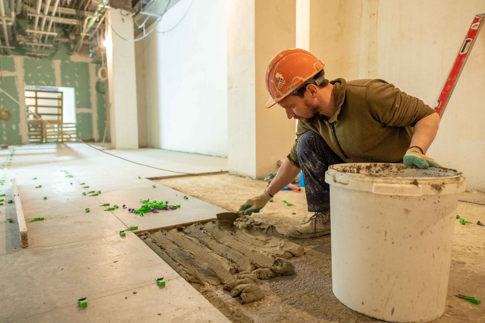 Un carreleur en casque de chantier étale du mortier-colle au peigne cranté sur le sol d'un local en cours d'aménagement intérieur, à côté des dalles grand format déjà posées.
