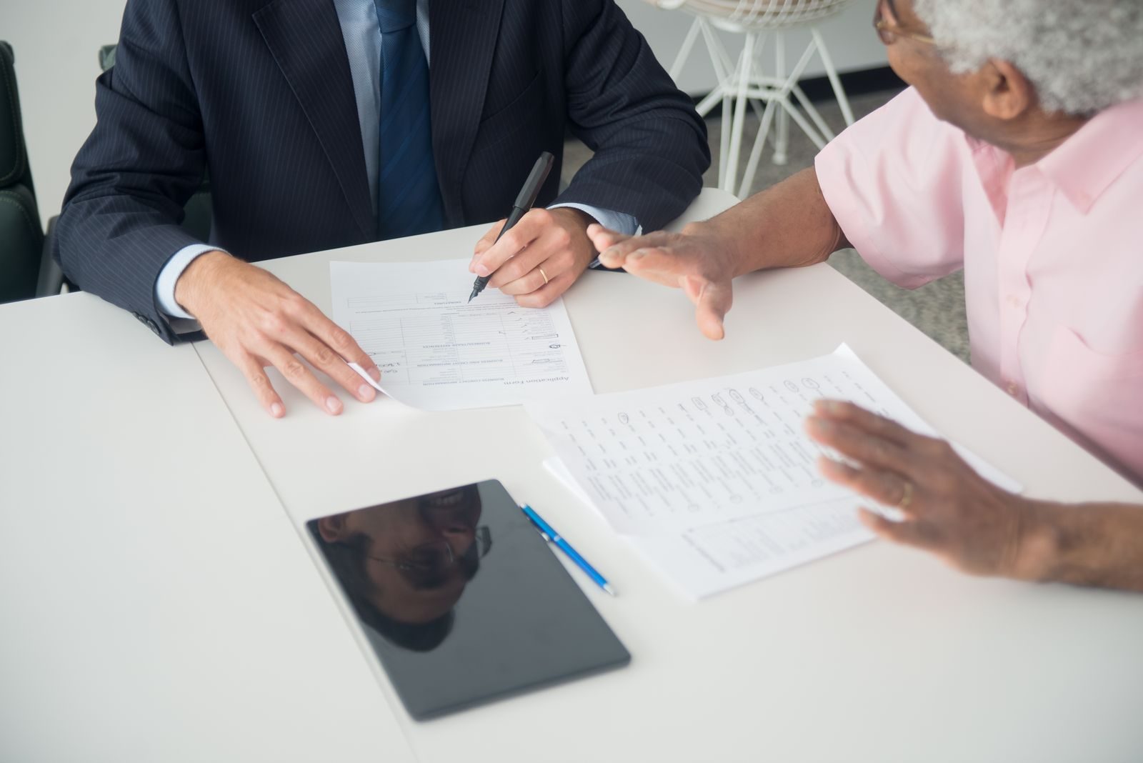 An adviser in a suit at a desk holds a pen over a form turned towards an older client, who is holding other documents and gesturing as he comments.
