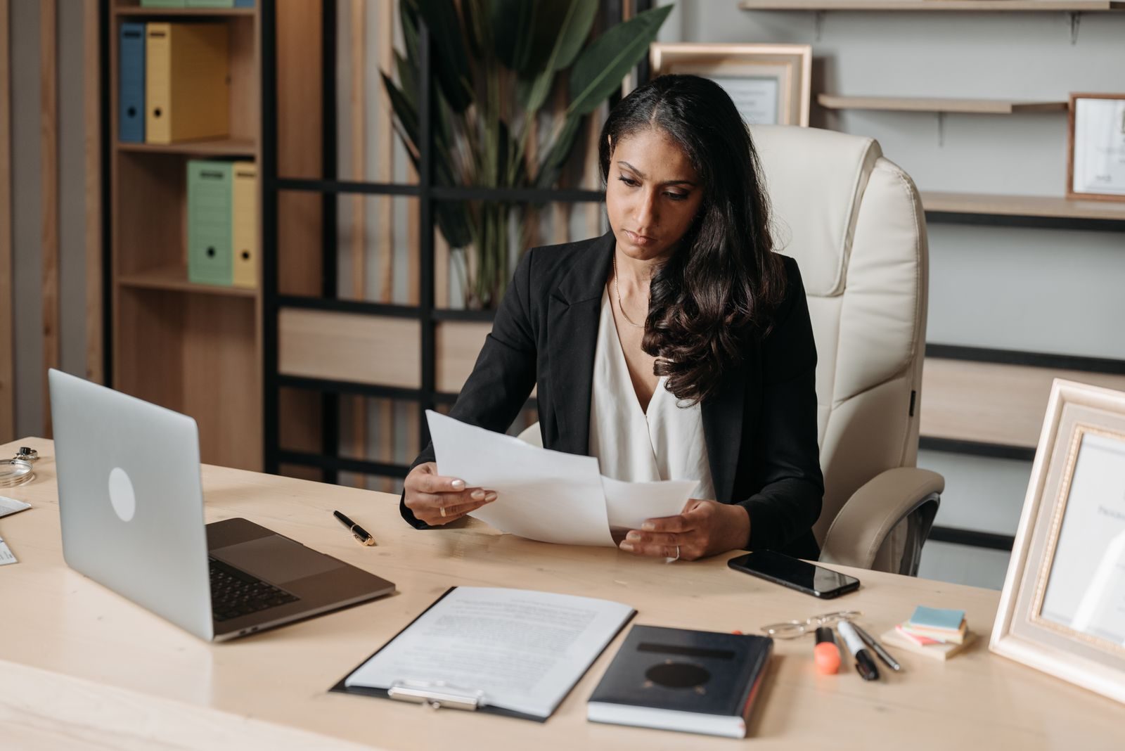 Une employée assise à son bureau lit deux documents papier ; devant elle un ordinateur portable et un porte-bloc, derrière elle une étagère garnie de classeurs d'archives.