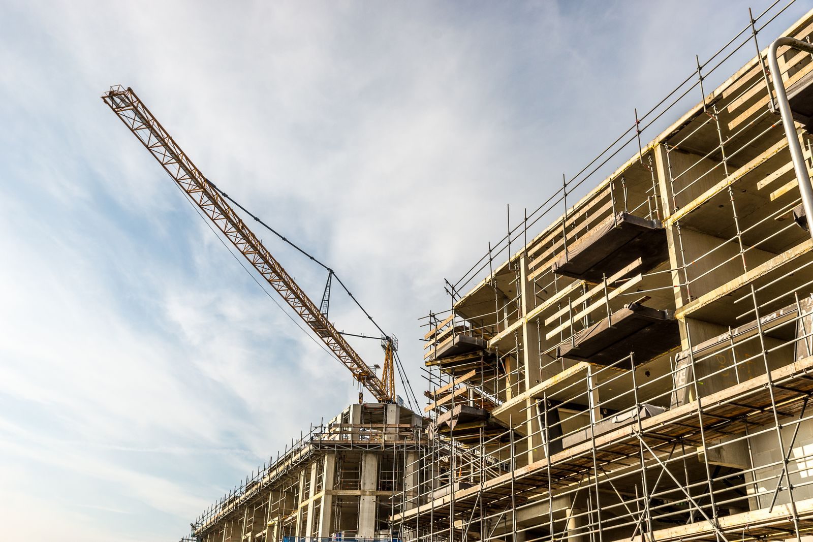 Un immeuble en construction vu d'en bas : sa structure de béton brut, sans façade, est entourée d'un échafaudage métallique, et la flèche d'une grue à tour jaune se détache sur un ciel bleu.
