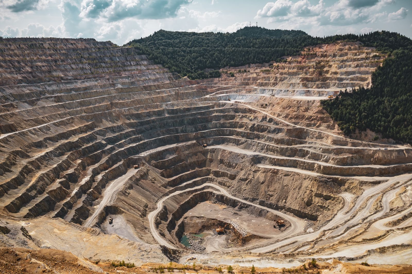 Vue en plongée d'une vaste carrière à ciel ouvert creusée en gradins étagés, entourée de forêt, avec des engins de chantier au fond de la fosse.
