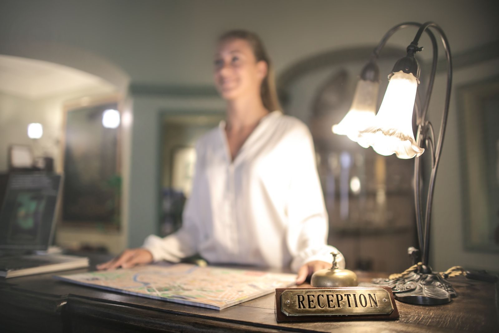 Le comptoir en bois d'une petite réception d'hôtel, avec une plaque en laiton « Reception », une sonnette de service et un plan de ville déplié ; derrière, une réceptionniste souriante.