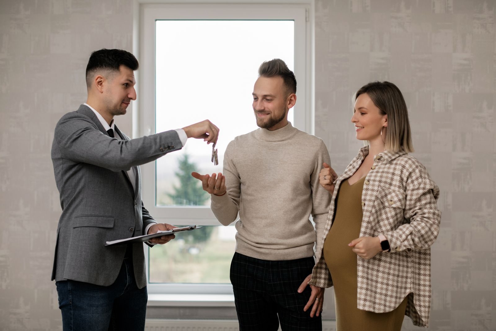 Un agent immobilier en costume remet un trousseau de clés à un homme, une femme enceinte à ses côtés, dans un appartement vide.