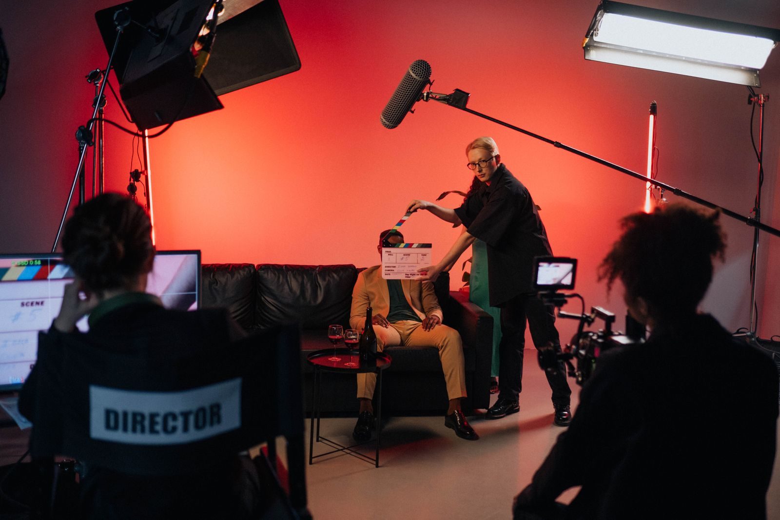 Sur un plateau de tournage éclairé en rouge, un membre de l'équipe tient un clap devant un comédien assis sur un canapé, pendant qu'une caméra et un moniteur de contrôle suivent la scène.