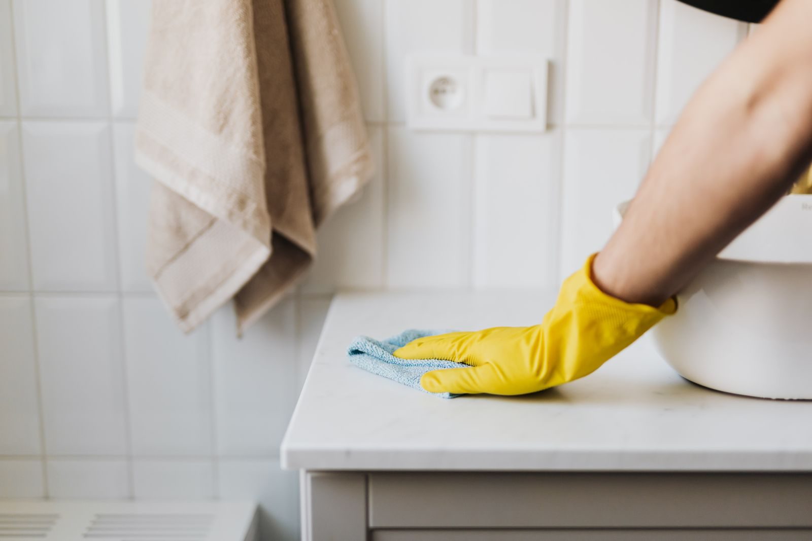 A hand in a yellow rubber glove wiping a white countertop with a microfibre cloth in a tiled bathroom.