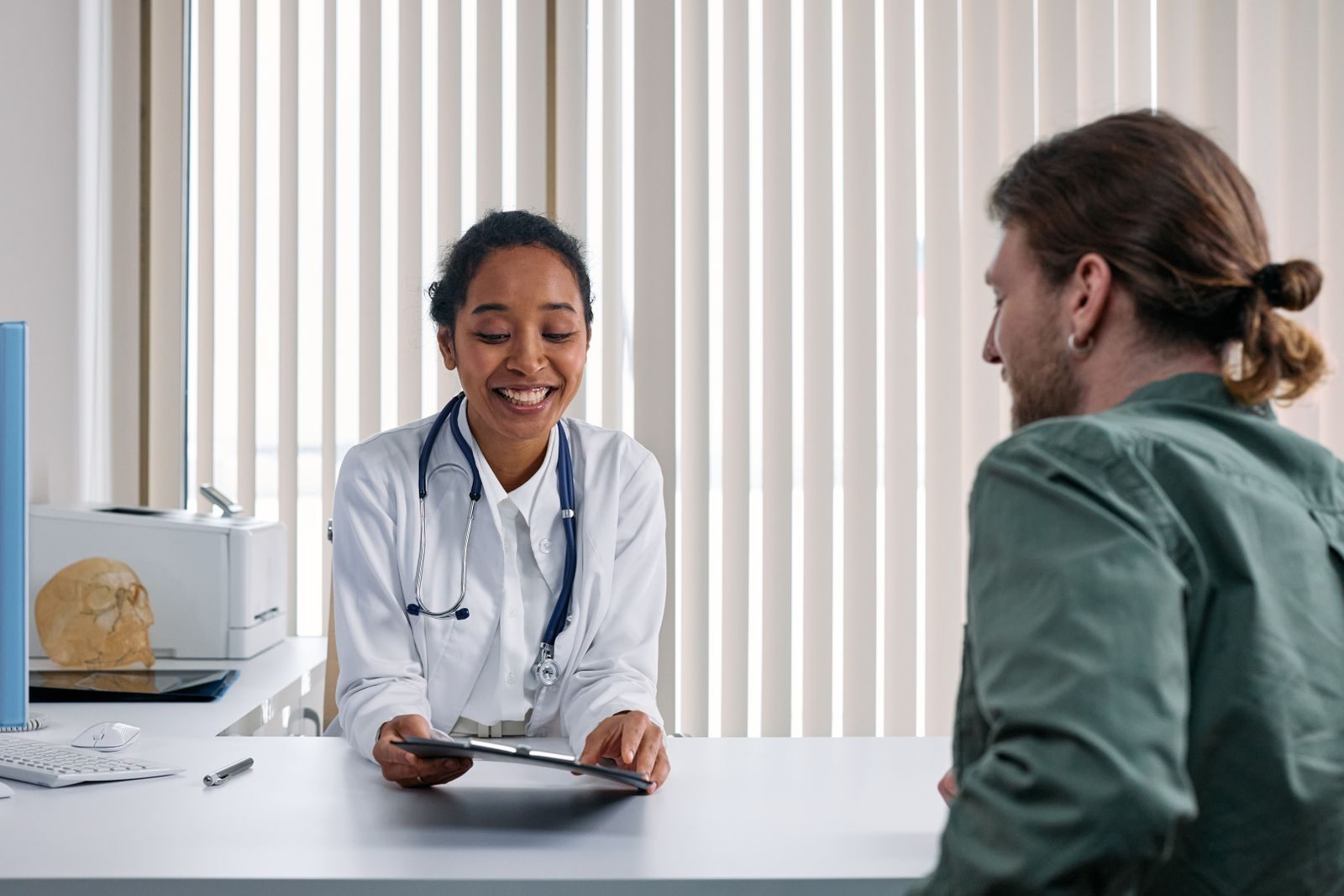 Une médecin en blouse blanche, stéthoscope autour du cou, présente une tablette à un patient assis en face d'elle dans une salle de consultation.