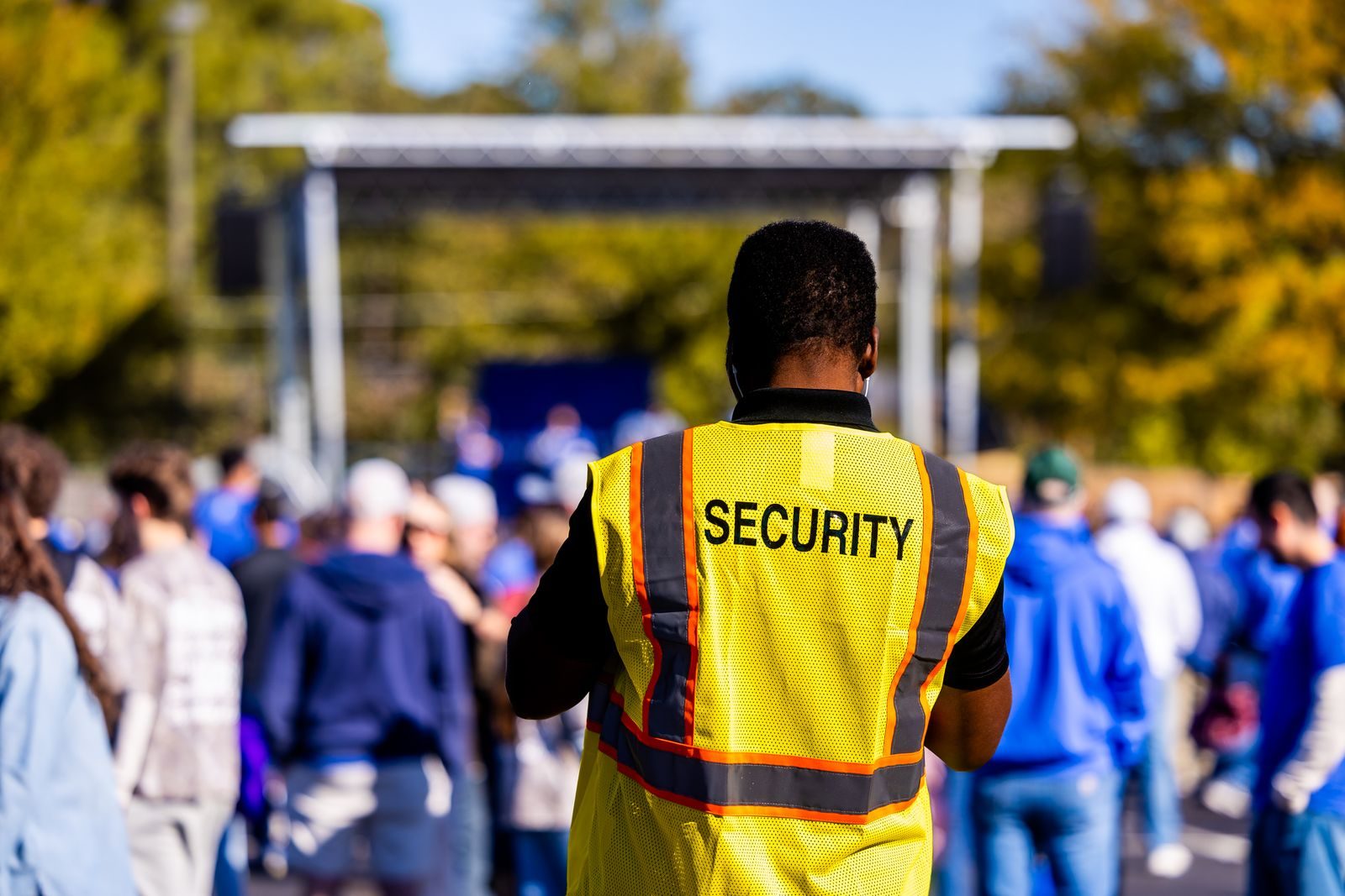 Un agent de sécurité en gilet jaune fluo floqué « Security » surveille de dos la foule rassemblée devant une scène, lors d'un événement en plein air.